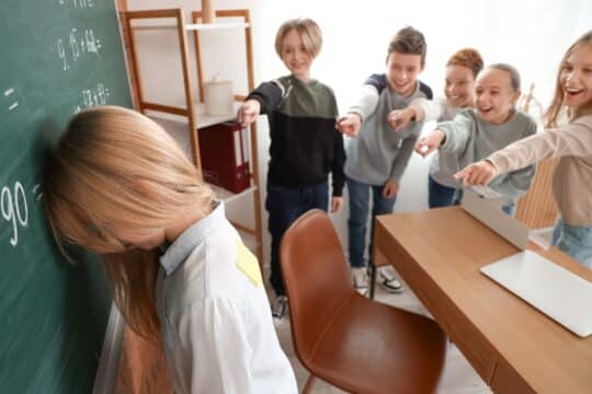 A group of students bully another student who stands at the chalkboard, hiding her face.