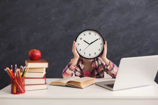 A student sits at their desk doing homework and holding a clock.