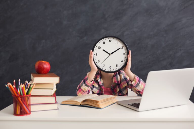 A student sits at their desk doing homework and holding a clock.