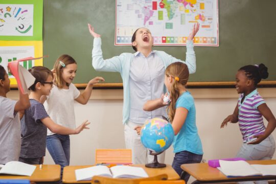 Students run around the classroom while a teacher holds her hands up in frustration.