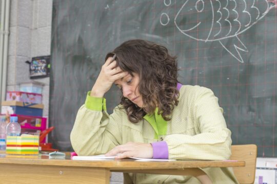 A stressed and exhausted teacher sits at her desk with her head in her hands, representing teacher burnout.