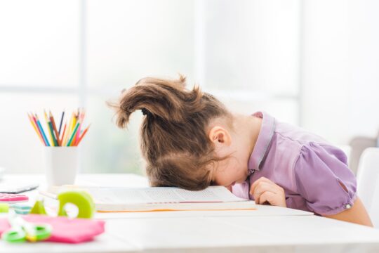 A young girl sits with her head down in her book, looking sad and frustrated.