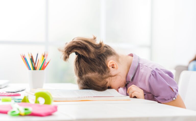 A young girl sits with her head down in her book, looking sad and frustrated.