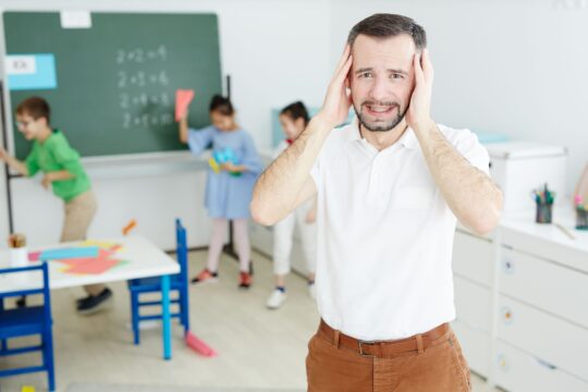 Portrait of a teacher standing with his hands on his head while his students run around the classroom.