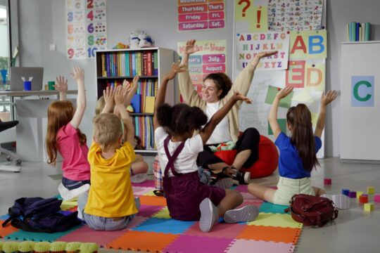 An elementary school teacher sits on the floor with her students, and they are moving their hands together in sync.