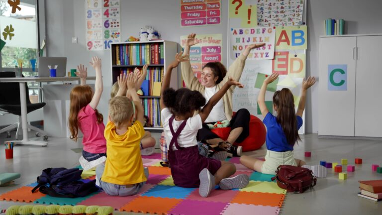 An elementary school teacher sits on the floor with her students, and they are moving their hands together in sync.