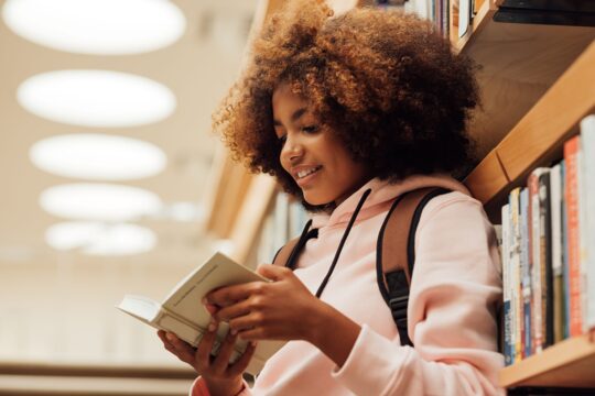 A student stands in the library, looking at a book she picked out.